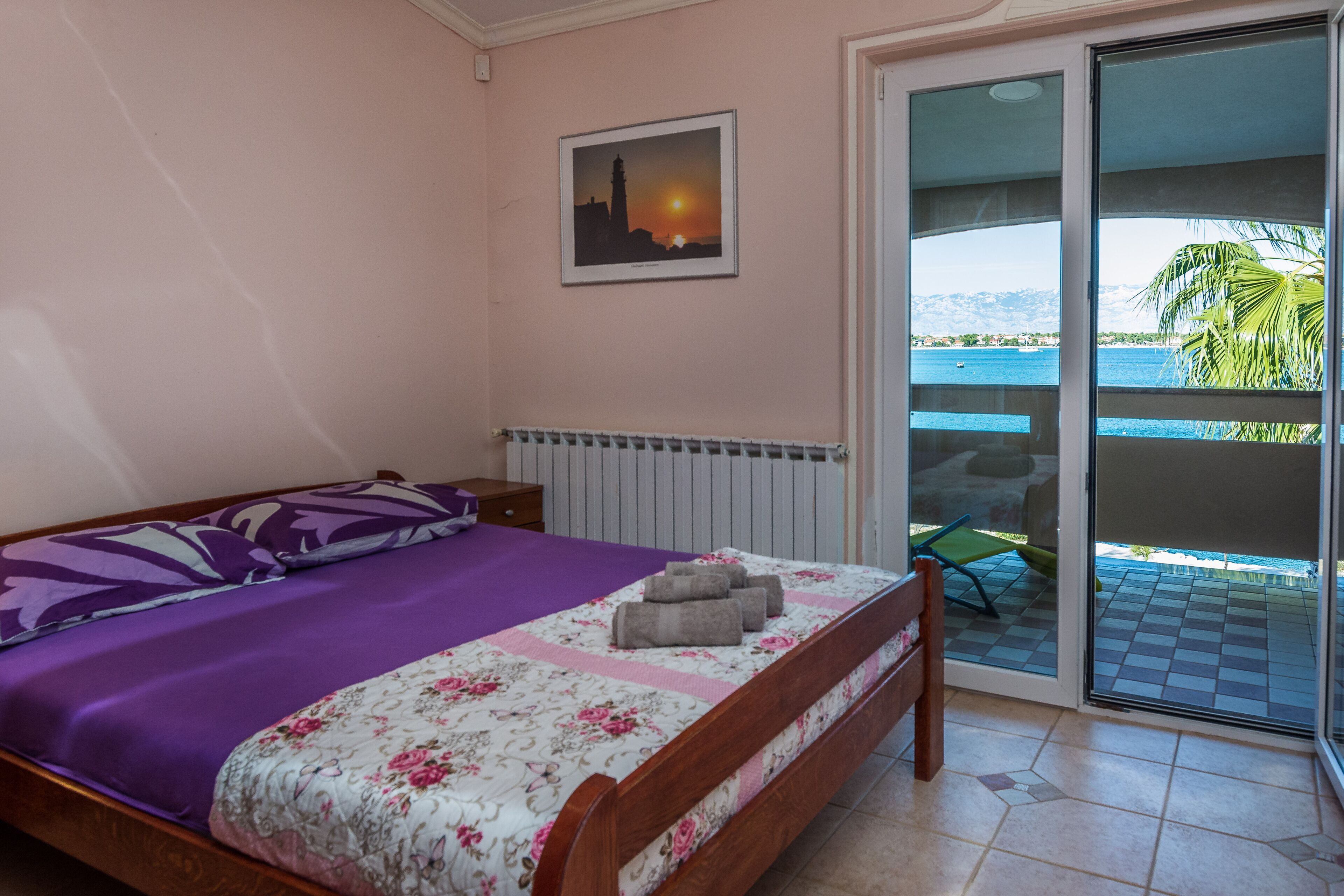 A bedroom with a bed, a window with a view of the sea, and a picture on the wall.