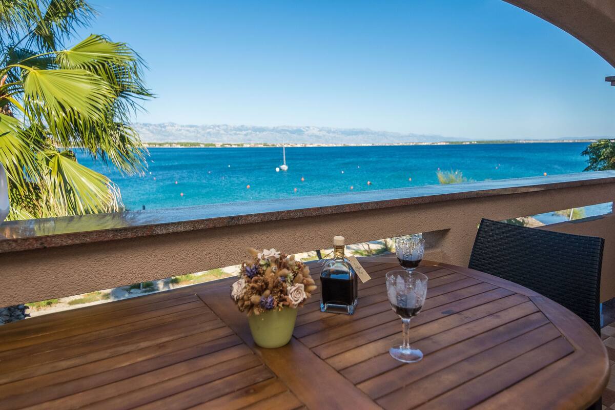 Una mesa de madera con una botella de licor, una copa y un ramo de flores, con vista a un mar azul claro y palmeras.