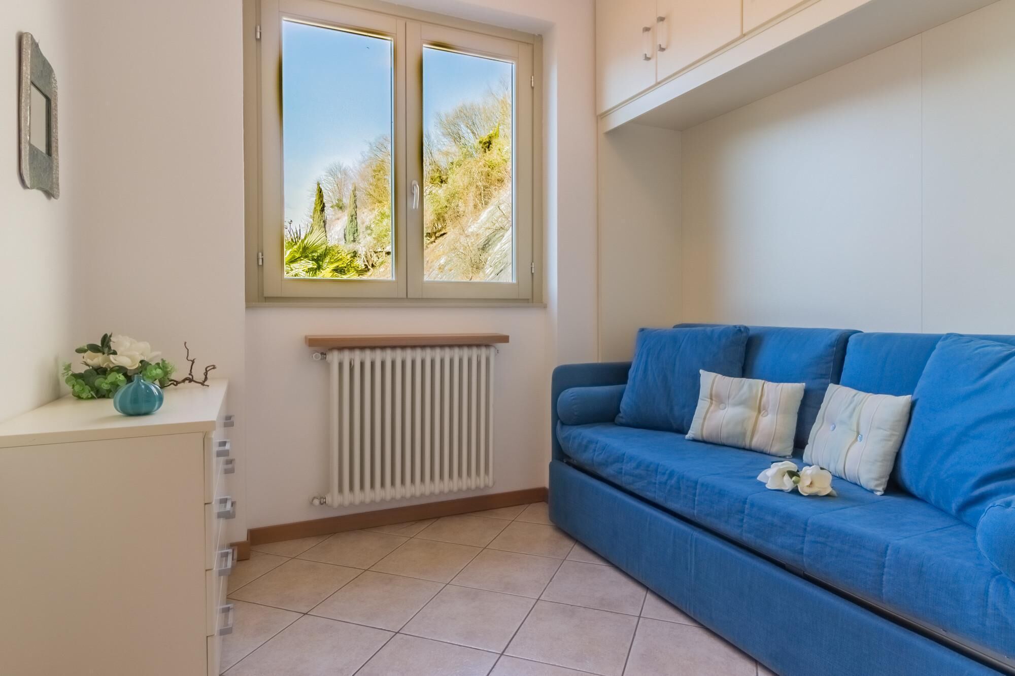 A room with a blue sofa, a radiator, a window with a view of trees, and a small cabinet with a vase of flowers.