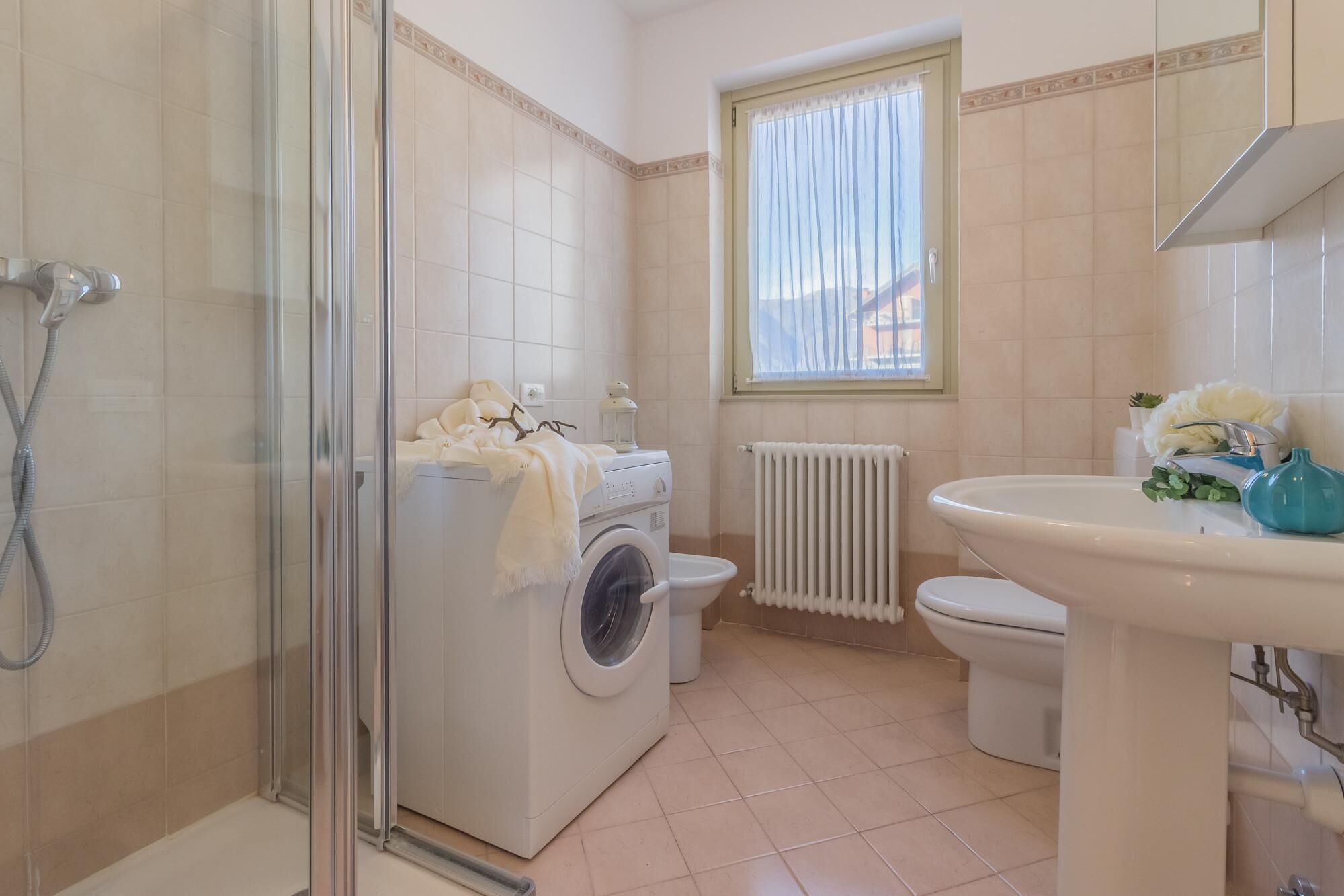 Uma casa de banho com chuveiro envidraçado, máquina de lavar roupa, sanita e lavatório com espelho em cima.