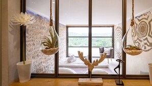 Une pièce avec une grande fenêtre, un lit recouvert de draps blancs, une chaise et deux pots de fleurs suspendus contenant des plantes.