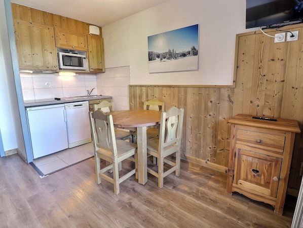Una cucina compatta con mobili in legno, un forno a microonde e un lavandino. C'è un tavolo da pranzo con sedie e un piccolo mobile in legno con un televisore.
