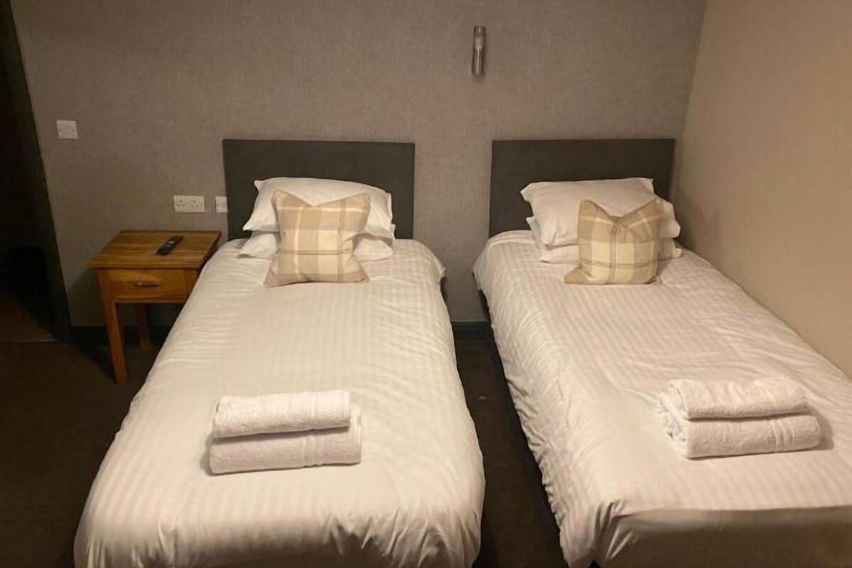 Two single beds with white linens and plaid pillows, a small wooden nightstand with a remote control, and a wall-mounted light fixture.