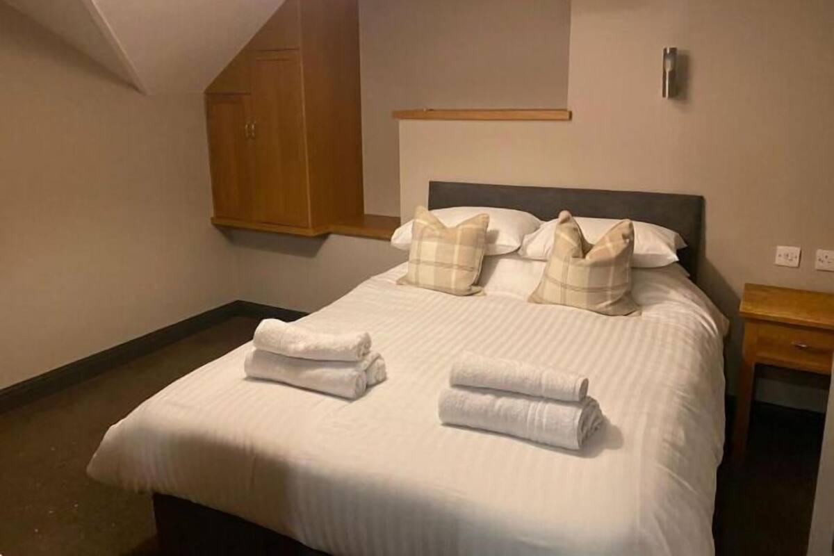 A neatly made bed with two rolled towels, a wooden wardrobe, and a bedside table with a lamp.