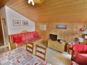 A cozy room with a wooden ceiling, a sofa with red cushions, a television on a wooden cabinet, and a table with a quilted tablecloth.