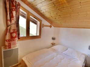 A bedroom with a wooden ceiling, a bed with white bedding, a window with curtains, and a wall-mounted lamp.