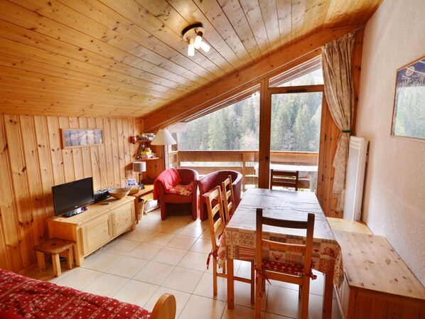 A cozy cabin with wooden walls and a large window overlooking a forest. It includes a dining table, chairs, a TV, and a small kitchen area.