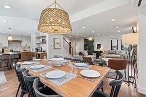 Un vaste espace de vie comprenant une table à manger en bois pouvant accueillir six personnes, un grand luminaire suspendu en fibres tressées et un escalier menant à un niveau supérieur.
