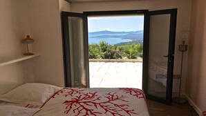Ein Schlafzimmer mit Bett, Nachttisch, Lampe und einer Glastür, die auf einen Balkon mit Blick auf das Meer und die Hügel führt.