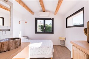 Un dormitorio con una cama, un banco de madera, una gran pila de piedra, una ventana y un ventilador de techo.