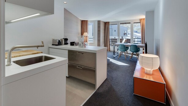 A modern hotel room with a kitchenette, a dining area with a table and chairs, and a view of the outside.