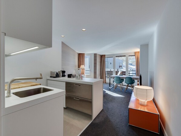 A modern hotel room with a kitchenette, a dining area with a table and chairs, and a view of the outside.