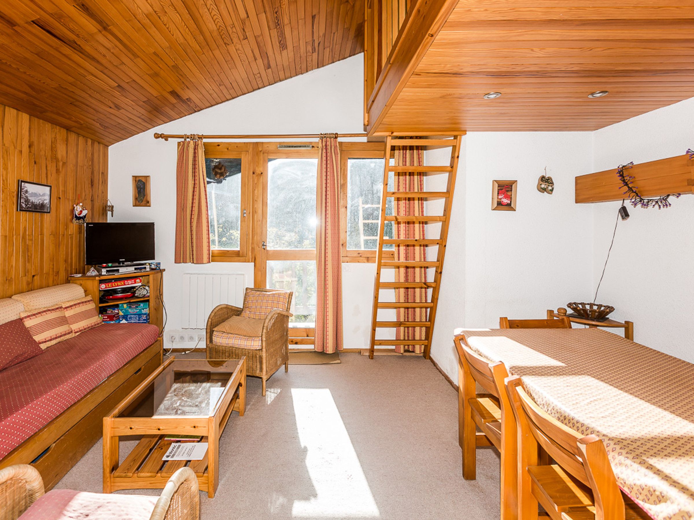 Una habitación acogedora con techo de madera, un sofá, un televisor, una mesa, una silla y una escalera.