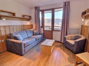 A cozy room with a sofa, armchair, wooden floor, and a view of mountains through a large window. - Comfortable 1-Bedroom, Ski-In/Ski-Out, Near Shops (La Tania)