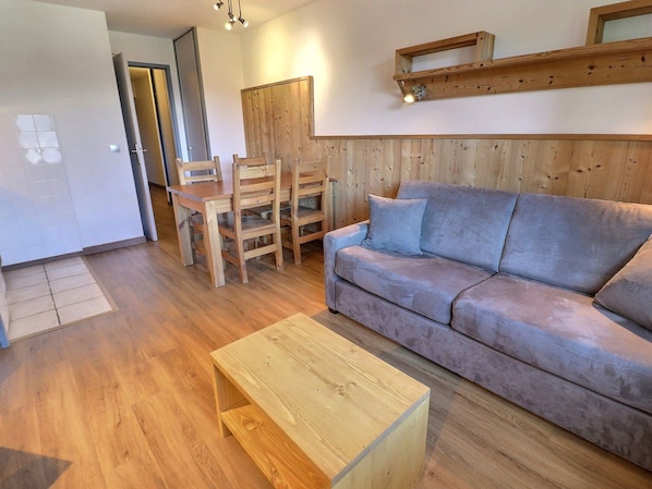 A living room with a wooden floor, a grey sofa, a wooden coffee table, and a dining area with wooden chairs and a table.