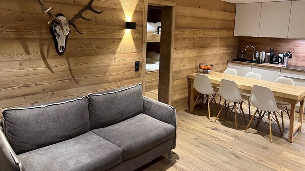 Een modern interieur met een houten wand, een opgehangen hertenschedel, een grijze bank en een eethoek met witte stoelen en een houten tafel.