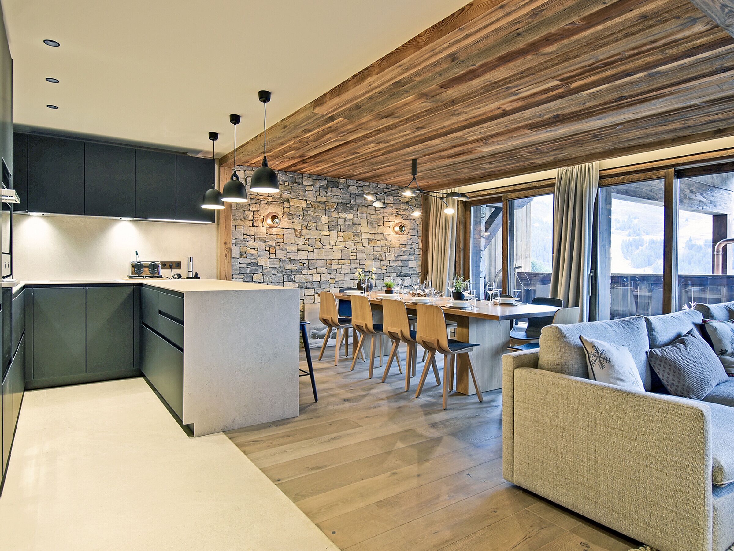 A modern kitchen with a stone wall, wooden ceiling, and a dining area with a large table and chairs.