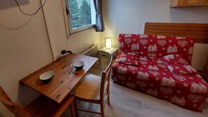 Ein kleines Zimmer mit Holztisch, einem Stuhl, einem Bett mit rotem, gemustertem Überwurf, einem Nachttisch mit Lampe und einem Fenster mit Blick auf Bäume.