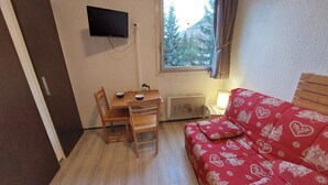 Ein kleiner Raum mit Schreibtisch aus Holz, zwei Stühlen, einem rot gemusterten Sofa, einem an der Wand montierten Fernseher und einem Fenster mit Blick auf Bäume und Gebäude.