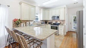 A kitchen with white cabinets, a granite countertop, a stainless steel refrigerator, and a dining area with wicker chairs. - Florida Cottage | Fenced yard| Serentiy Cottage | Hot Tub | Santa Rosa Beach (Santa Rosa Beach)