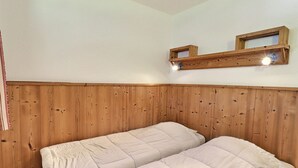A room with wooden paneling on the walls, two beds with white bedding, and a shelf with two light fixtures. - Comfortable 2-Room, Ski-In/Ski-Out, Close to Shops (La Tania)
