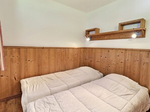 Une pièce aux murs recouverts de lambris en bois, avec deux lits dotés de draps blancs et une étagère sur laquelle sont posés deux luminaires.