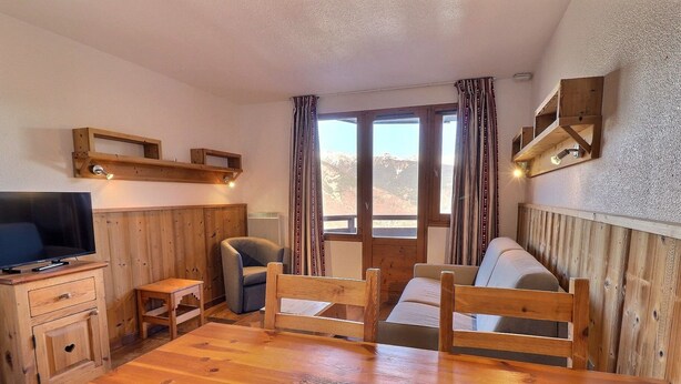 A cozy room with a wooden dining table, a sofa, a TV, and a window with curtains. - Comfortable 2-Room, Ski-In/Ski-Out, Close to Shops (La Tania)