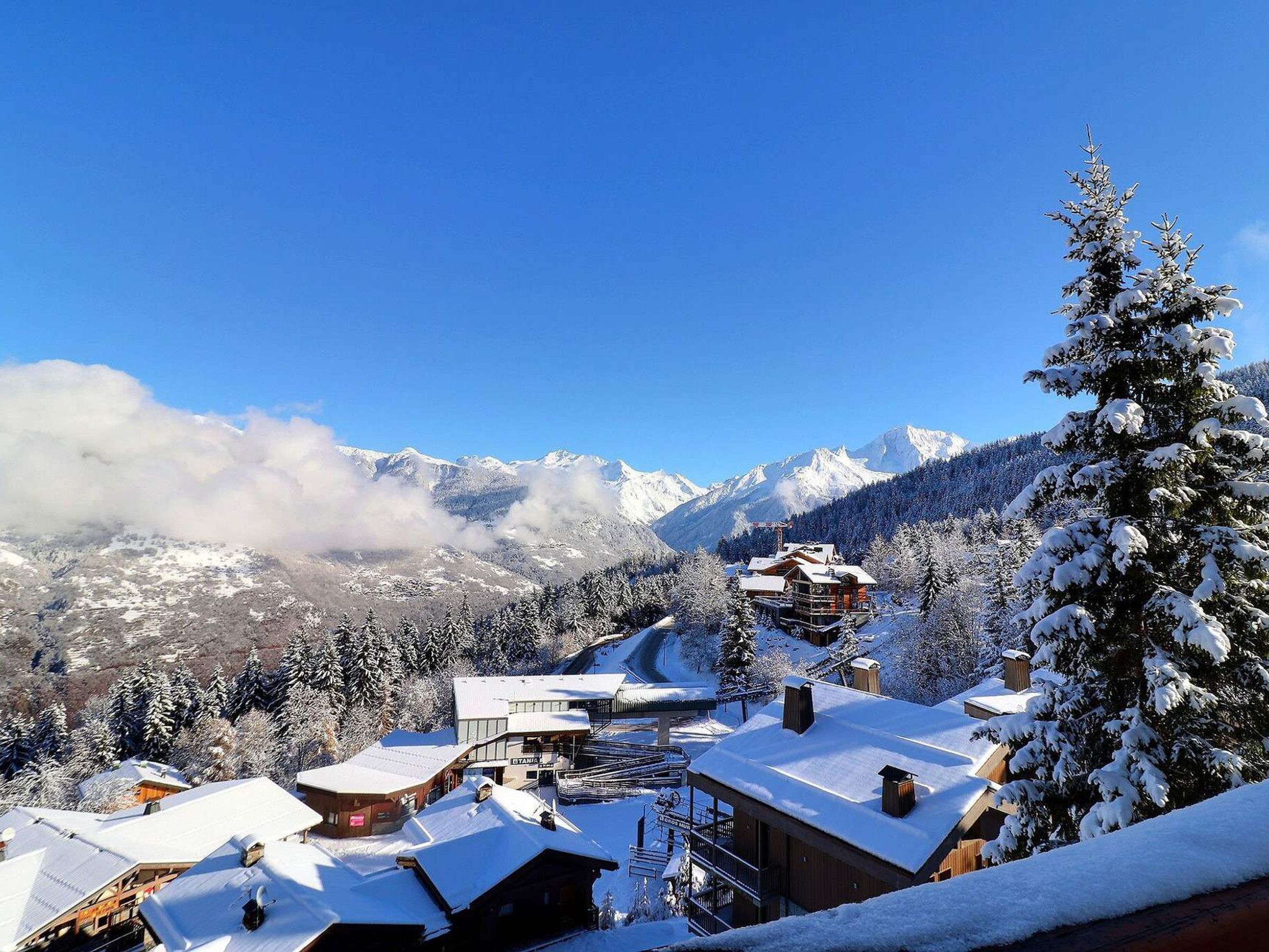 A snow-covered mountain resort with wooden buildings, a ski lift, and a clear view of snow-capped peaks.