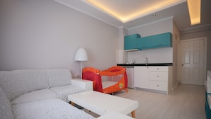 Un salon moderne avec un canapé, une petite table et un parc pour enfants. On aperçoit, au fond, une cuisine avec des armoires de couleur turquoise et des appareils électroménagers blancs.