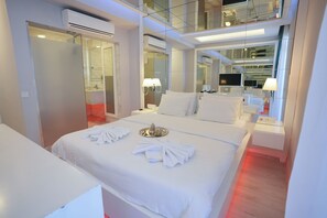 Una habitación de hotel moderna con una cama grande, una mesita de noche con lámpara, televisor y espejo.