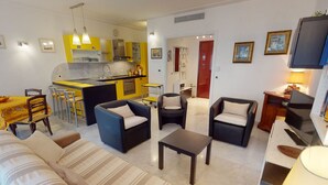 A modern living room with a sofa, armchairs, and a coffee table. A kitchen with yellow cabinets and a dining area with chairs is visible in the background. - Spacious 1-bedroom with AC, terrace and private garage near the sea (Menton)