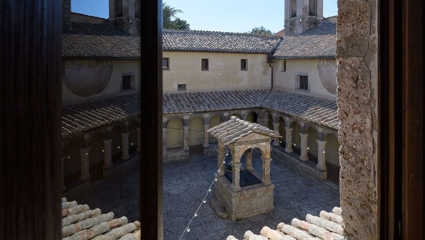 A courtyard with stone walls and a central fountain, viewed through a window with wooden frames. - Convento dei Cappuccini (Tolfa)