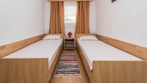 A small, clean room with two single beds, a wooden headboard, a small table with a lamp, and a window with curtains. - Apartment Bubanovic (Vir)
