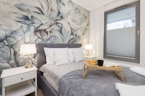 Une chambre avec un grand décor mural floral, un lit gris, des oreillers blancs, deux lampes de chevet, un plateau avec un livre et du jus d’orange, et une fenêtre avec des stores.