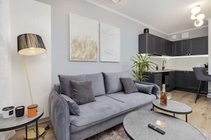 Un salon moderne avec un canapé gris, une table basse ronde et un lampadaire. On y trouve également un coin cuisine avec des meubles sombres et un tabouret de bar.
