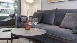 A modern living room with a grey sofa, a round coffee table, a bottle of wine, two wine glasses, a remote control, and a floor lamp.