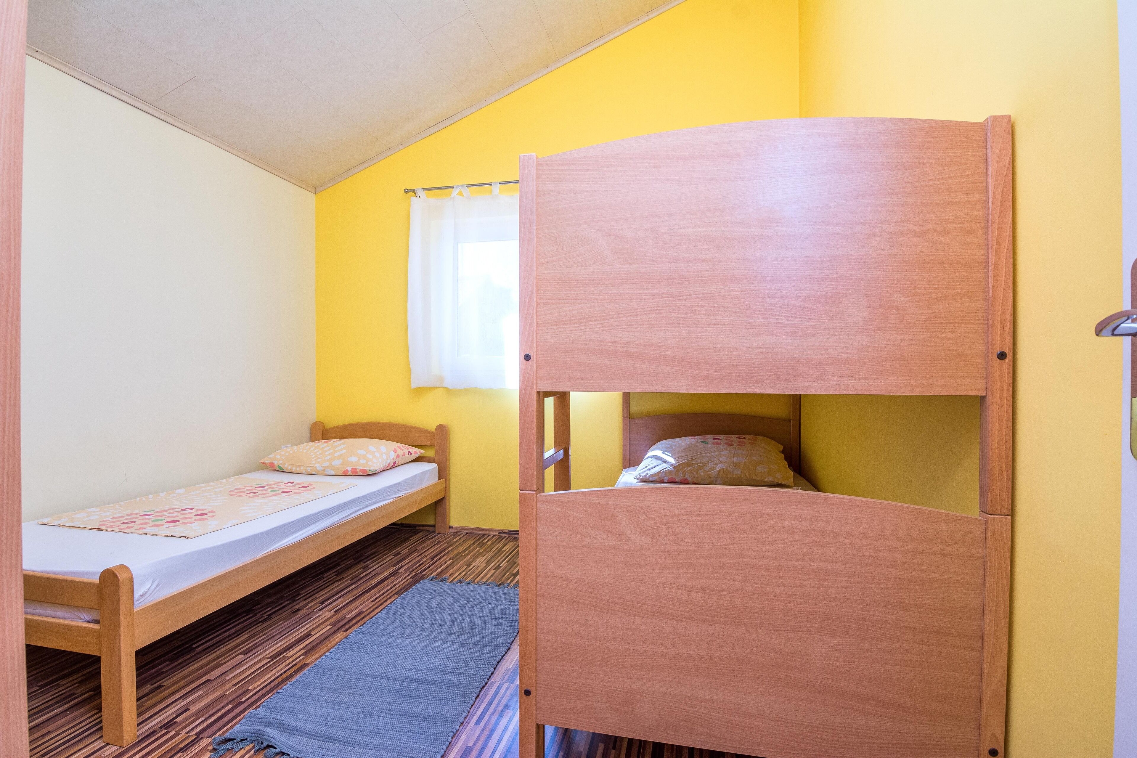 A room with a single bed, a bunk bed, a window with curtains, and a wooden floor.