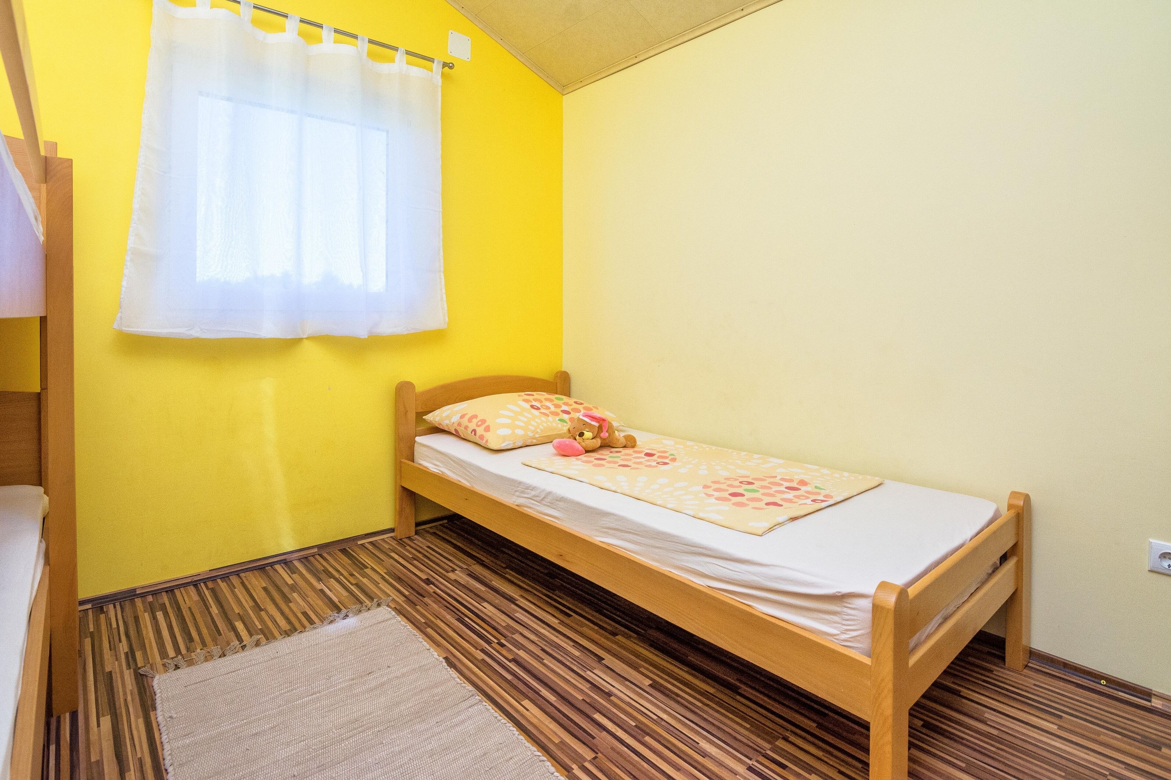 A small, single-bed room with a wooden frame, a yellow wall, a white curtain, and a patterned bedspread.