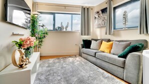 A modern living room with a grey sofa, a grey rug, a white side table with a vase of flowers, and a large window with curtains. - Tower Bridge Modern 2Bed 2Bath Apartment by London Bridge (London)