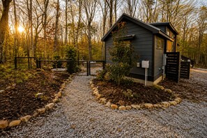 Une petite cabane, accessible par un chemin de gravier, entourée d’une clôture et d’arbres.