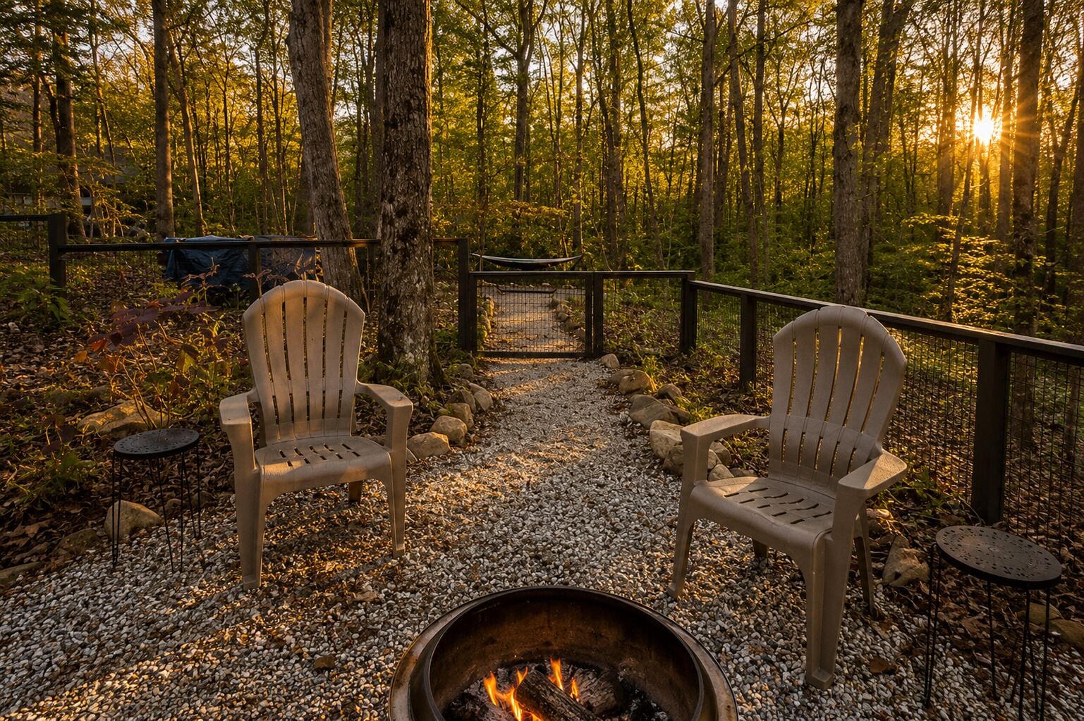 Deux fauteuils Adirondack sont placés face à un foyer, dans un espace boisé où l’on trouve un chemin de gravier et une clôture en bois.