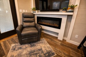 Een leren fauteuil voor een open haard, met een televisie erboven, een vloerkleed en een raam met zonwering.