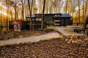 A cabin with a deck, surrounded by trees and a gravel path.