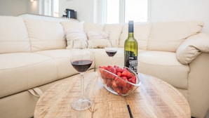 Una mesa de madera redonda con un tazón de fresas, dos copas de vino y una botella de vino en una sala de estar con un sofá color beige y un ventanal grande.