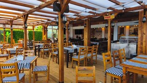 Ein Restaurant mit Terrassensitzgelegenheiten, Holztischen und -stühlen sowie einem überdachten Bereich. Schilder weisen auf Fisch- und Pizzen.