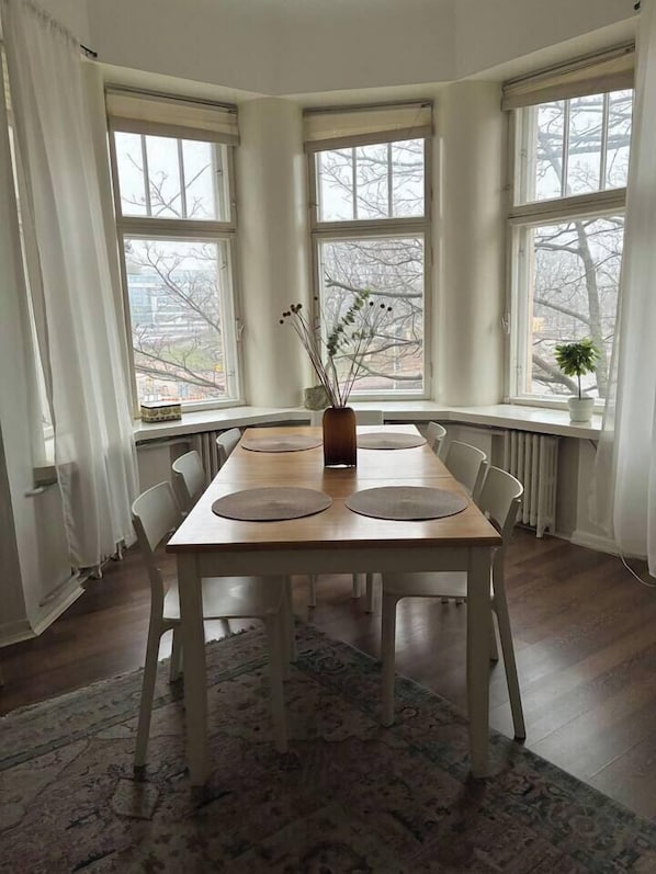 A dining area with a wooden table, white chairs, a central vase with plants, and large windows with curtains.