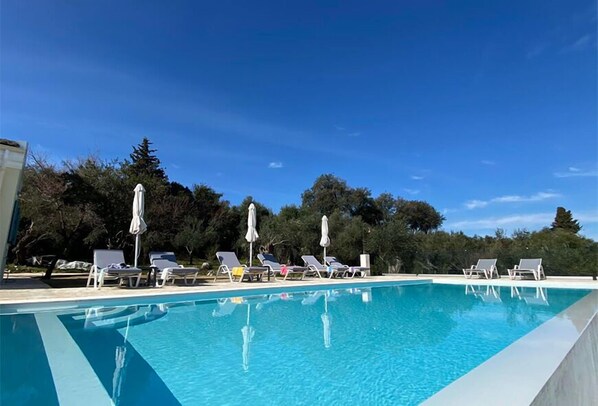 A clear blue swimming pool with lounge chairs and umbrellas, surrounded by trees and a clear sky.
