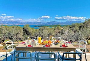 An outdoor dining table with chairs, set with plates, cutlery, and a fruit bowl, overlooking a scenic landscape with mountains and a body of water.