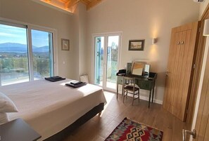 A bedroom with a large bed, a desk, and a wardrobe. There are large windows offering a view of the outdoors.