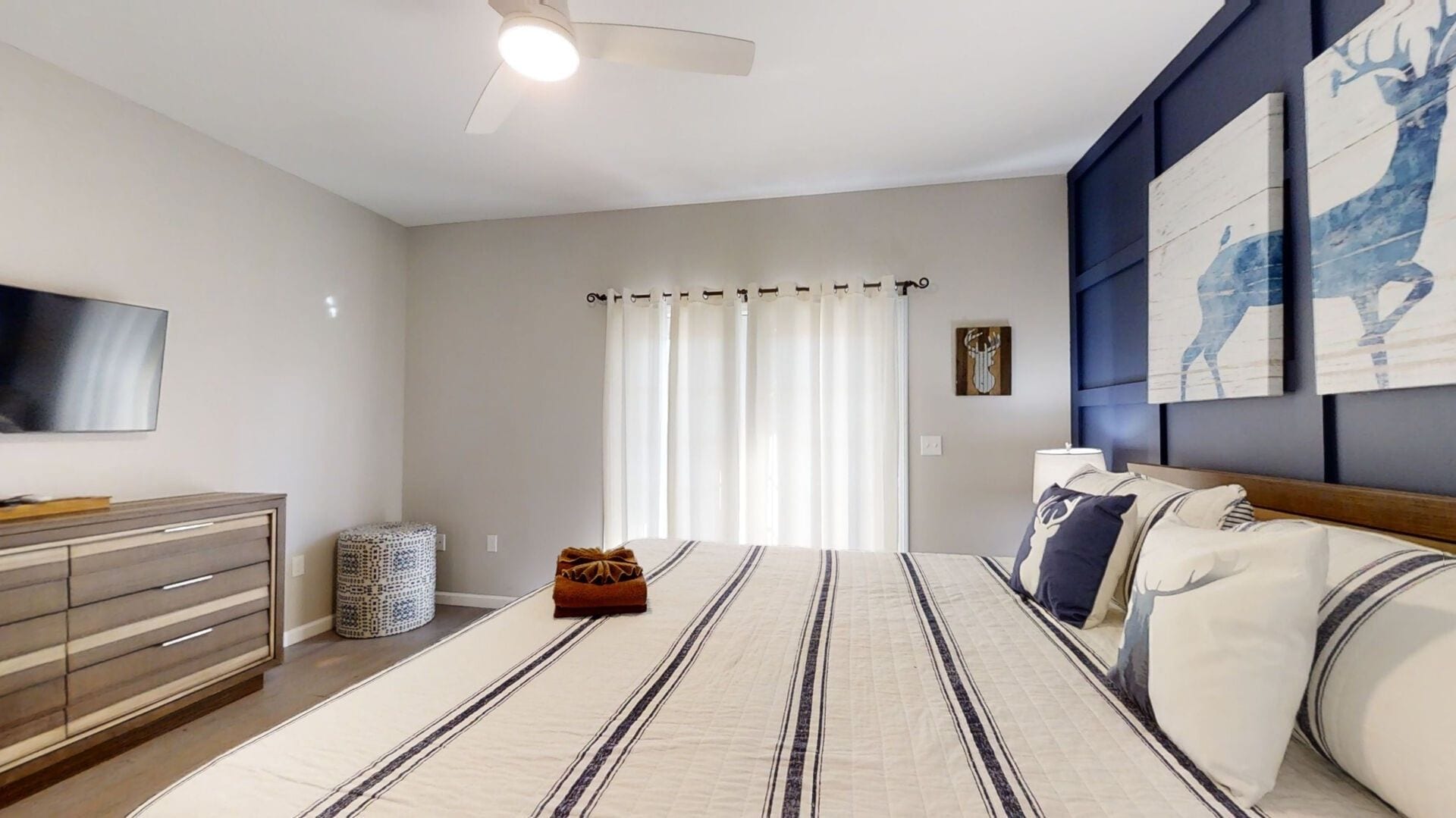 Un dormitorio con cama, televisor, ventilador de techo, ventana con cortinas y una obra de arte en la pared.